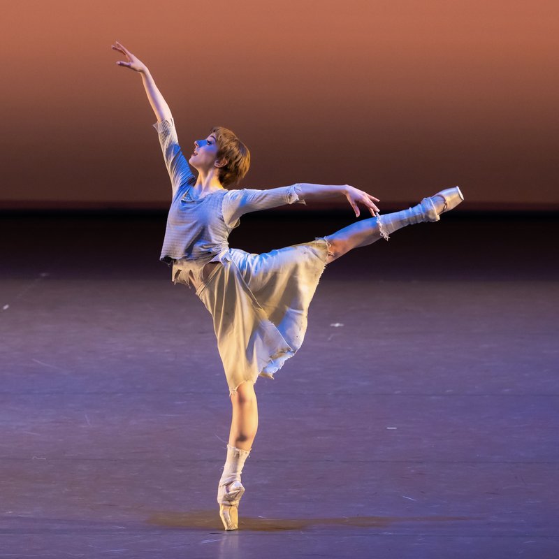 A female presenting dancer in a tattered white skirt and layered grey top steps onto pointe in an arabesque, eyes tilted up towards her hand.