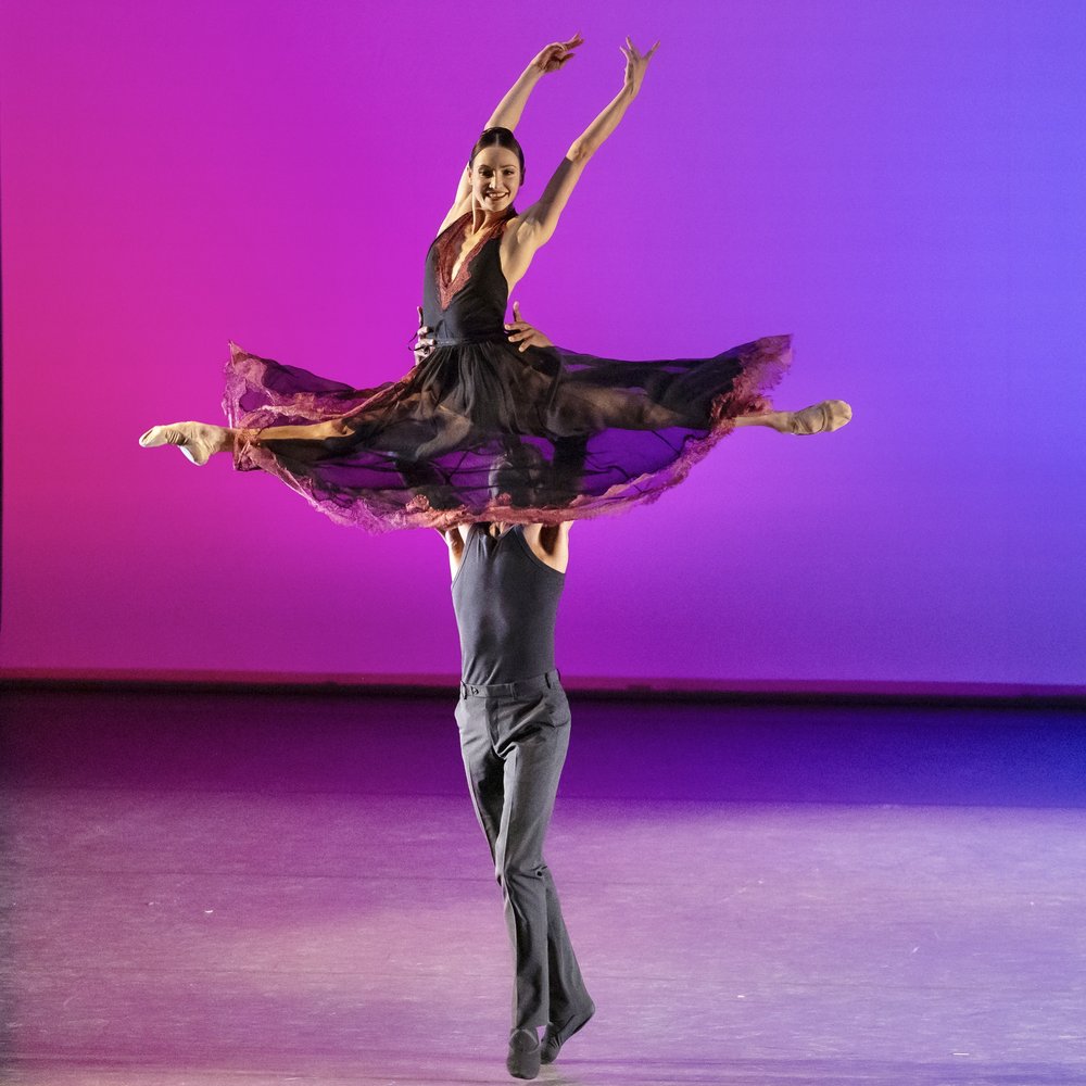 On a stage in front of a pink, blue, and purple gradient backdrop, a male presenting dancer in black pants and a black tank top lifts a female-presenting dancer above his head into a split. Her skirt, sheer black with a magenta lace edge, flies out.