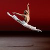 A male presenting dancer springs into a 180 degree split in the air with their arms lifted in an L. Their bare chest, white pants, and white socks stand out against the red backdrop of the stage.