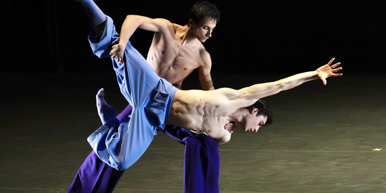 Two male presenting dancers, with their chests exposed and wearing matching flowy blue pants and socks, engage in a dynamic lift with one dancer balanced on the other's knee. Luke Kwo / LK Studio photo.
