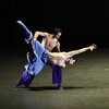 Two male presenting dancers, with their chests exposed and wearing matching flowy blue pants and socks, engage in a dynamic lift with one dancer balanced on the other's knee. Luke Kwo / LK Studio photo.