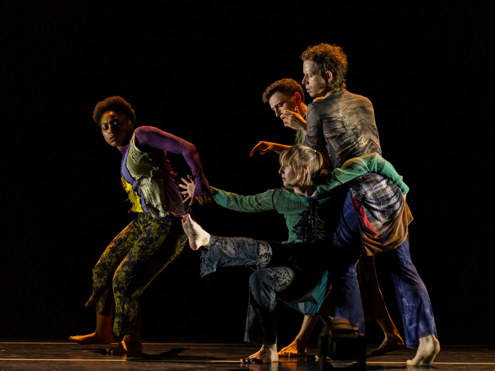 Four dancers create a connected pose onstage, with one seated low to the ground and reaching forward while the others stand and lean around them.