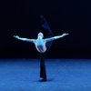 A female presenting dancer is tinged in blue light on a stage with a black backdrop. The dancer kicks their black-clothed leg like a scorpion tail above their head, arms open wide and head tilted back, so that their face is fully up to the ceiling.