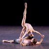 A female presenting dancer in a nude-colored top and shorts is partnering with a male presenting dancer with a bare chest and grey pants. She arches back with one leg in the air, hands supported by the male dancer on the ground, and they make eye contact.