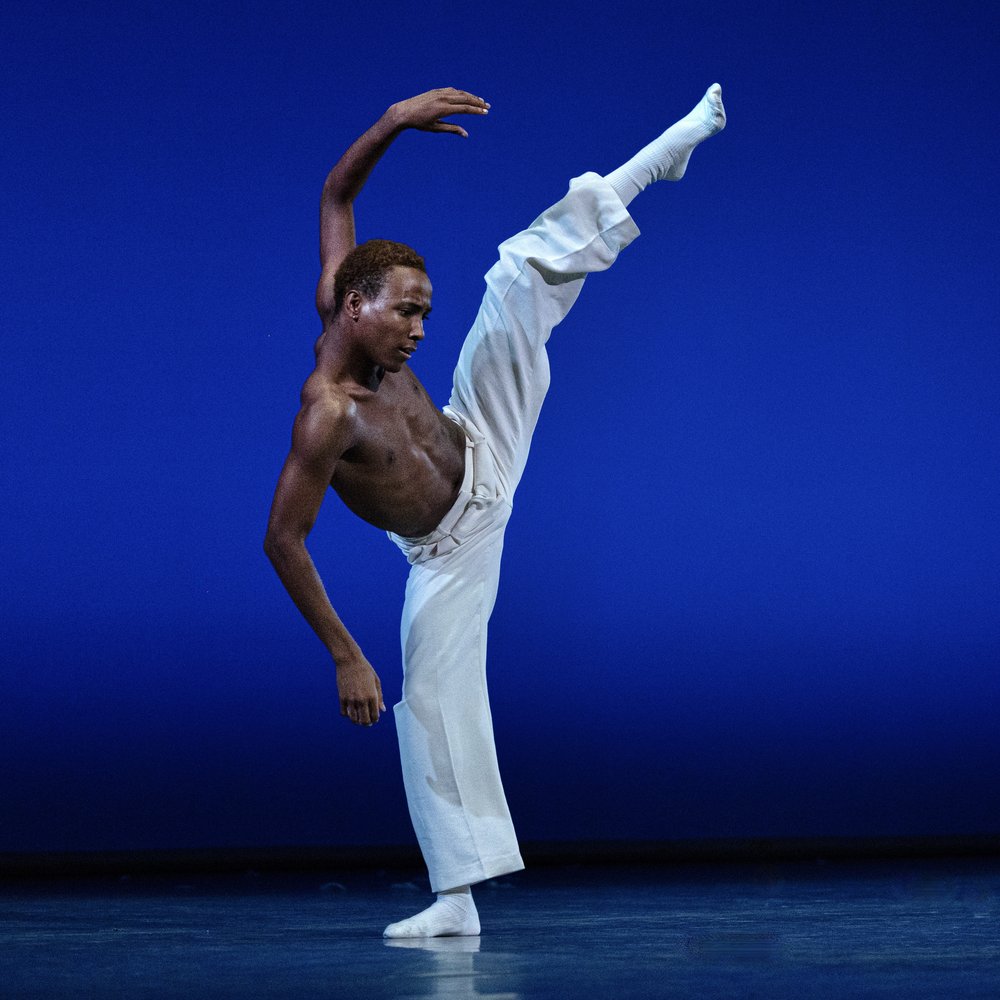 A Black male presenting dancer, with their chest exposed and wearing white wide pants and white socks, kicks his slightly-bent leg into the air in a split, matching the shape with lightly bent arms, on stage in front of a blue backdrop.