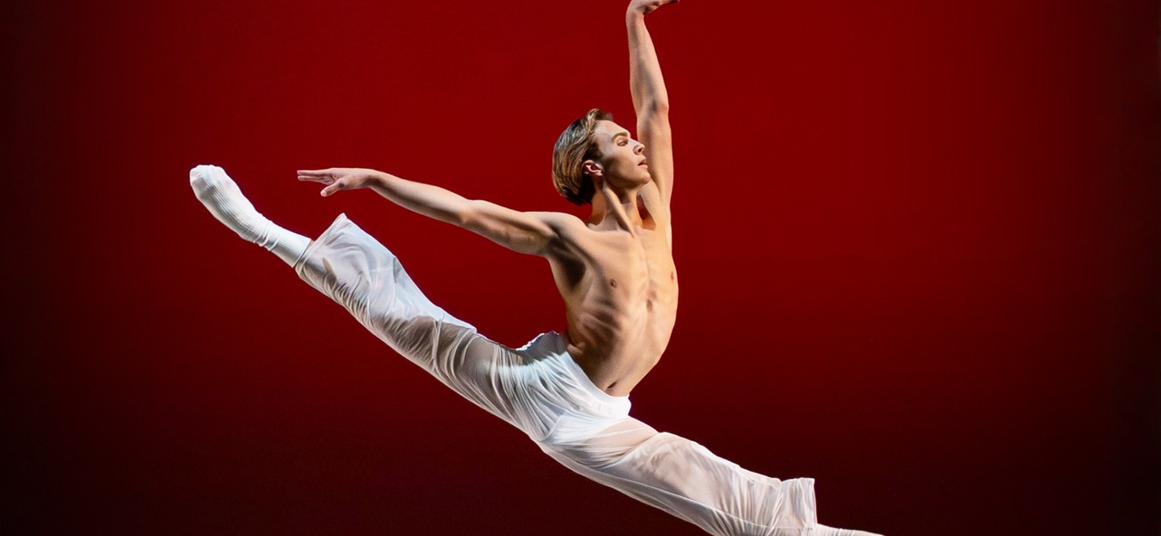 A male presenting dancer springs into a 180 degree split in the air with their arms lifted in an L. Their bare chest, white pants, and white socks stand out against the red backdrop of the stage. Luke Kwo / LK Studio photo.