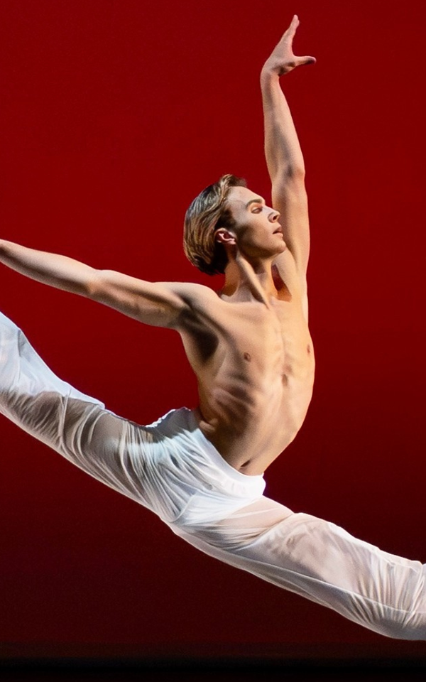 A male presenting dancer springs into a 180 degree split in the air with their arms lifted in an L. Their bare chest, white pants, and white socks stand out against the red backdrop of the stage. Luke Kwo / LK Studio photo.