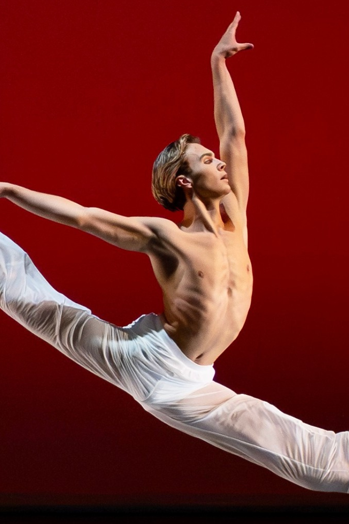 A male presenting dancer springs into a 180 degree split in the air with their arms lifted in an L. Their bare chest, white pants, and white socks stand out against the red backdrop of the stage. Luke Kwo / LK Studio photo.