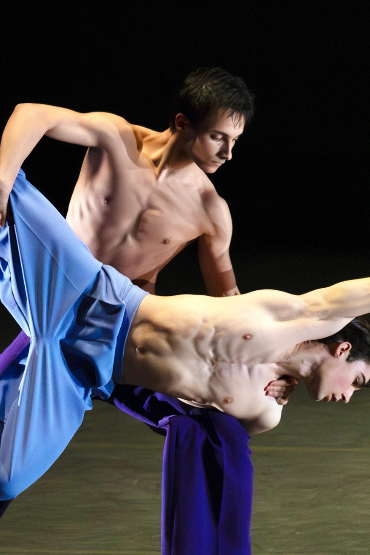 Two male presenting dancers, with their chests exposed and wearing matching flowy blue pants and socks, engage in a dynamic lift with one dancer balanced on the other's knee.