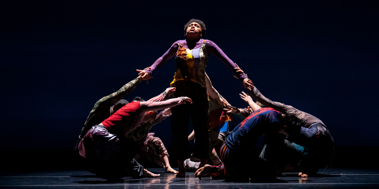 A group of dancers, in mis-matched clothes, are crouched on the ground, circling one Black dancer standing upright. The dancer's face is tilted up to the ceiling, yet their arms are being pulled down by the grabbing hands of the rest of the group.
