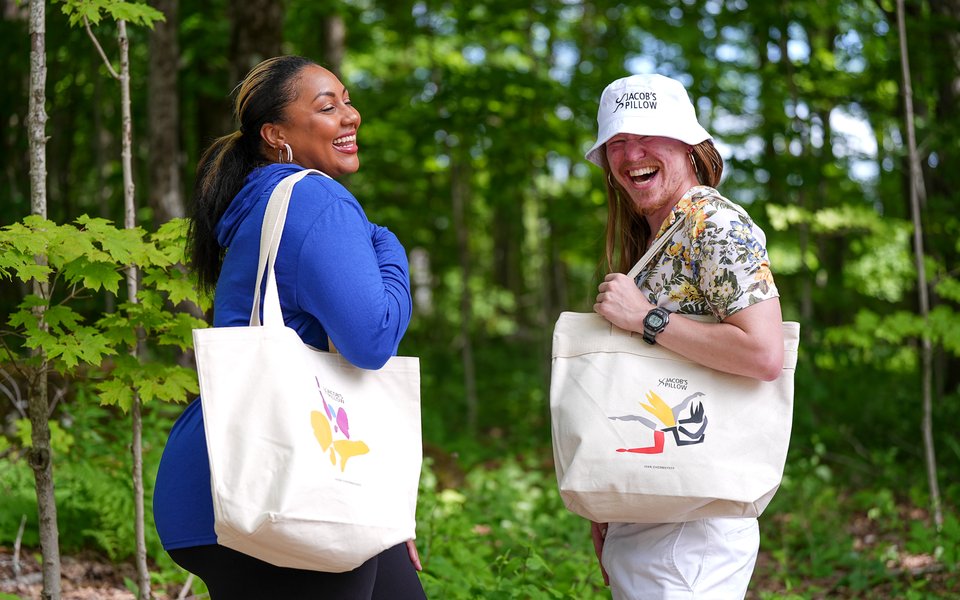 Two people facing one another, laughing, and holding Jacob's Pillow totes bags to their sides.