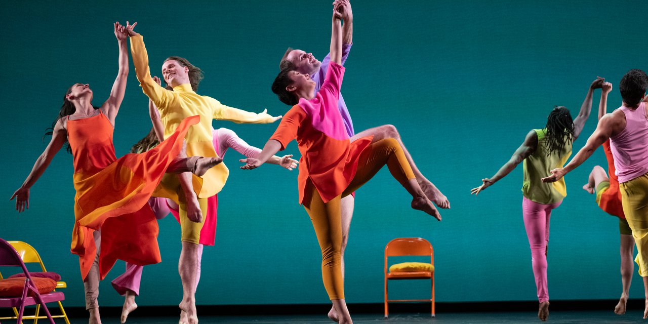 Coupled dancers jumping high in the air with hands lifted and intertwined. The dancers wear brightly colored, mismatched garments.