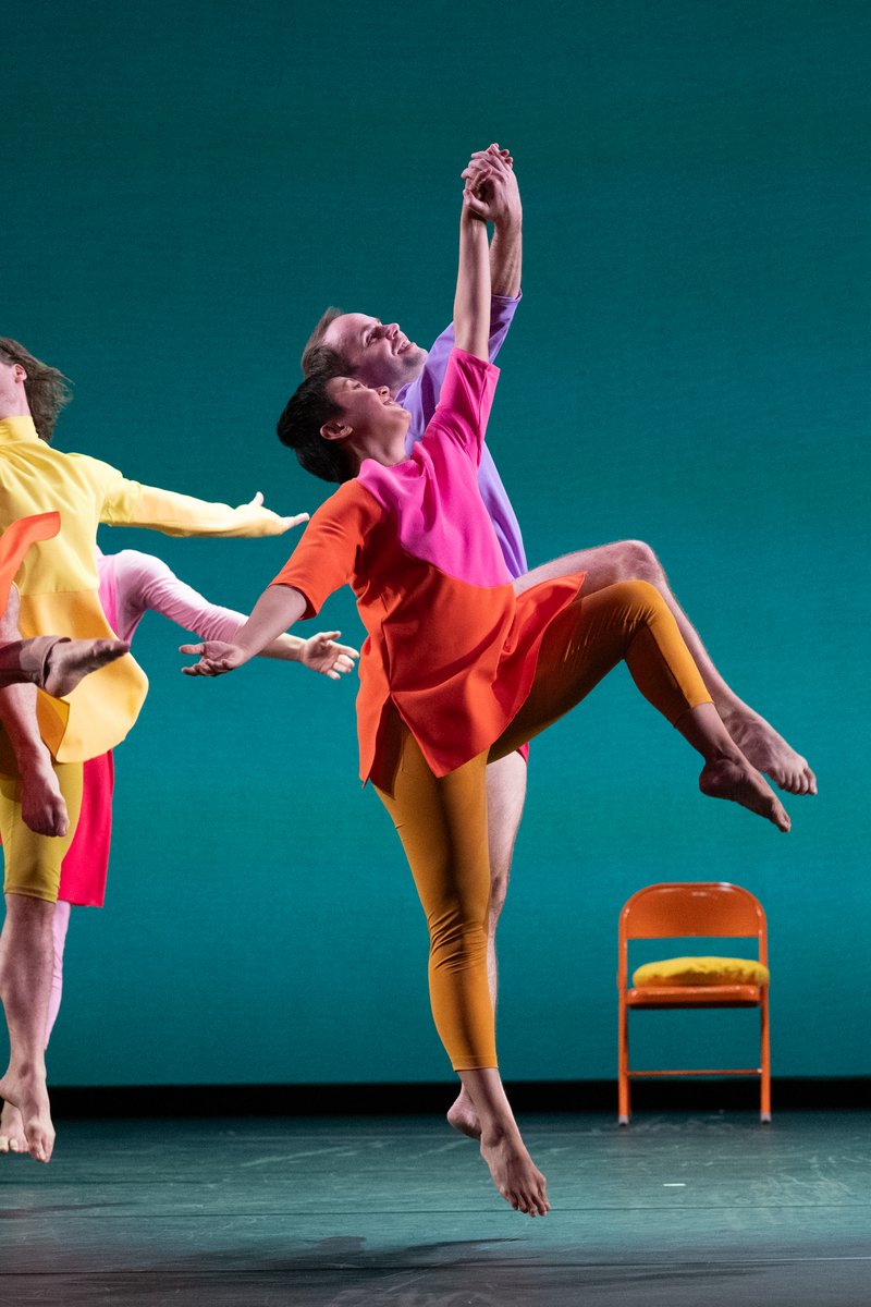 Coupled dancers jumping high in the air with hands lifted and intertwined. The dancers wear brightly colored, mismatched garments.