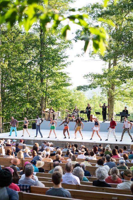 A number of performing artists line the front of the side facing the audience, legs in a wide position. A band plays on a second tier of the outdoor stage, backdropped and surrounded by green foliage.