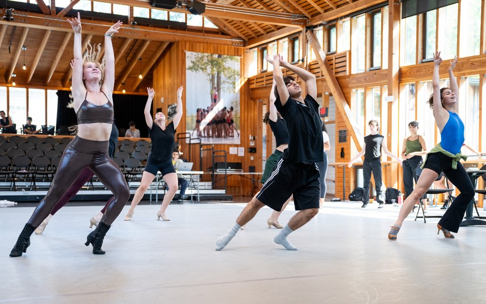 Three dancers stand on their left leg pointing their right leg to the side. they extend both arms straight above their heads and look upwards.