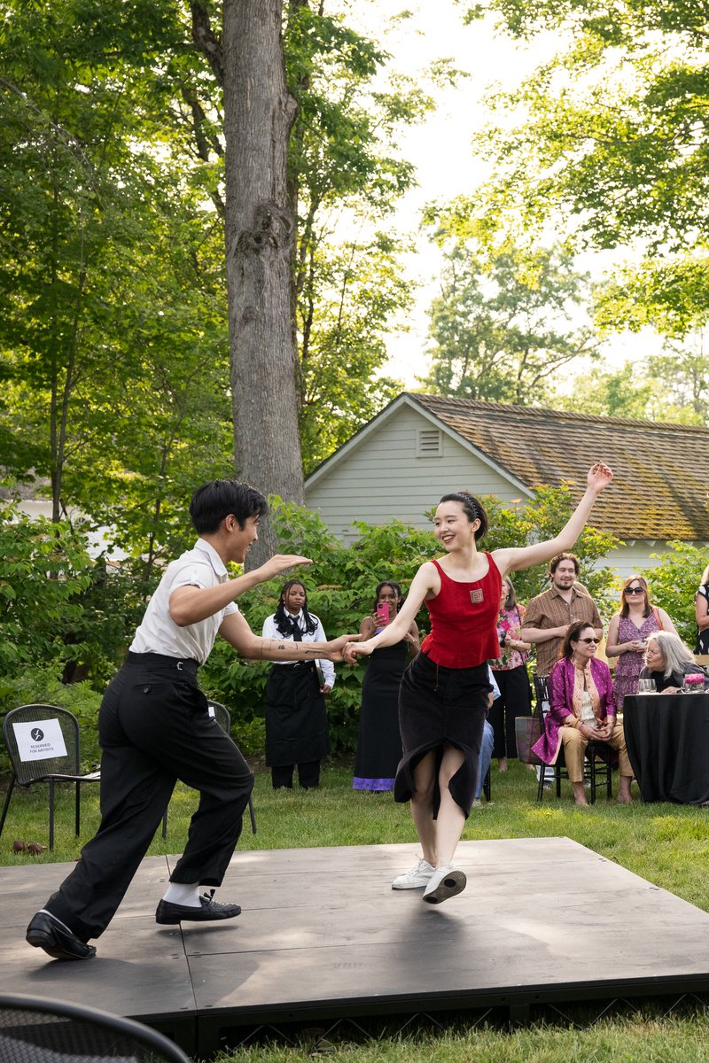 Two dancers in the middle of a partnered swing dance smile at one another. They are on a raised temporary stage. A large crowd of patrons on a great lawn watch intently.