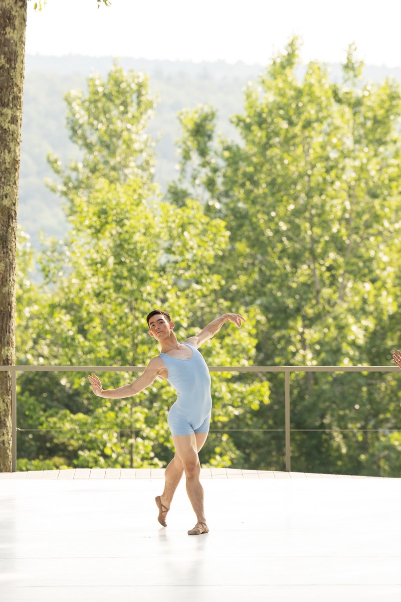 Three Dancers of The School lined up, arms in a diagonal and one leg reaching back. They are performing on the outdoor stage which features a lively green foliage backdrop.