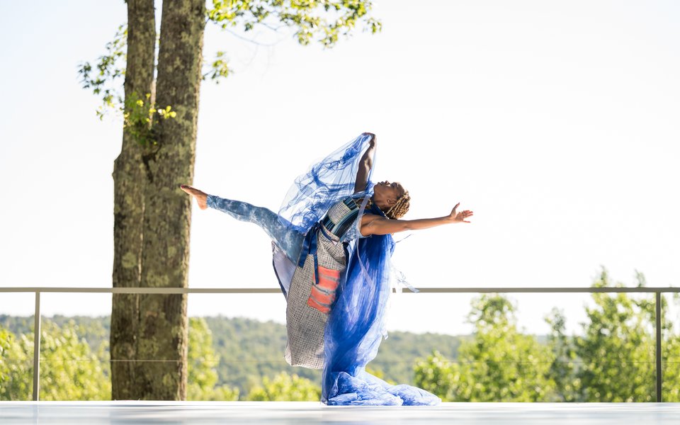 A performer leans back on one leg as the other extends up and out, chest up. Against the outdoor stage's lively green hilled backdrop, the dancer wears a vibrant blue flowing fabric.