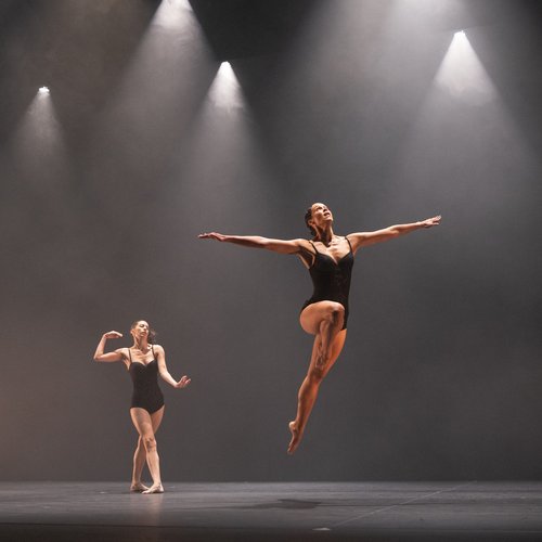 Two dancers in black leotards are lit with five cones of light on a black hazy stage. One is gesturing with their arms, the other leaping through the air, arms outstretched, one leg extended underneath them.