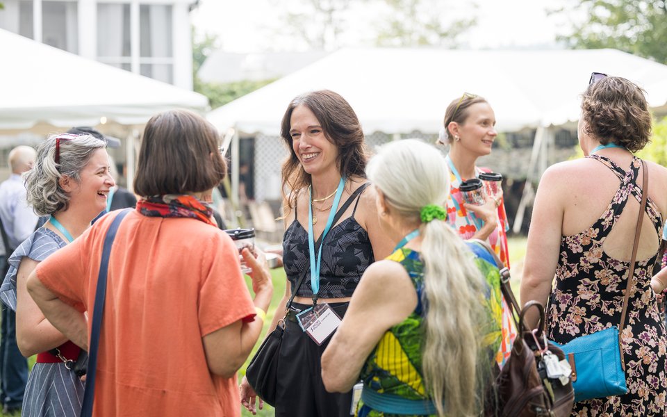 People mingle on a lawn in front of event tents. One person facing the camera and wearing a blue lanyard talks to three other attendees.