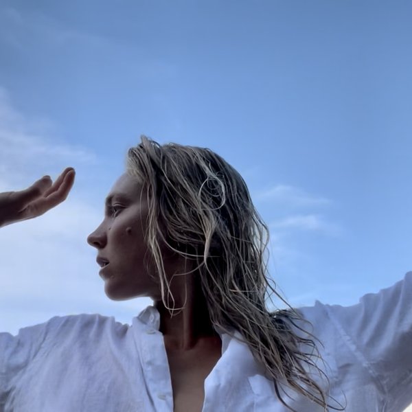 Image of a female-presenting dancer with blonde hair wearing a white shirt and raising their arms toward the sky.