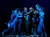Seven dancers gather in a loose circle onstage, some embracing or holding one another while a dancer stands at the center facing the audience.