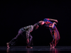 Two dancers perform onstage against a black background, with one lunging forward and the other leaning backward. Their heads touching, supporting each other's weight.