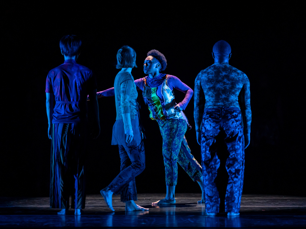 Four dancers are spaced across the stage under blue light, with one dancer facing another at center while two others stand with their backs to the audience.