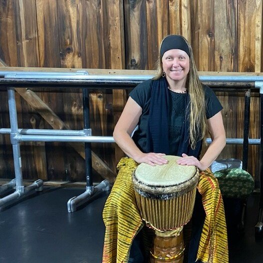 A woman with long hair sits with an African drum at her feet and smiles for the camera.