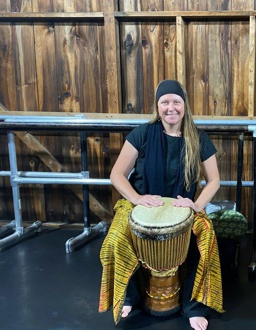 A woman with long hair sits with an African drum at her feet and smiles for the camera.
