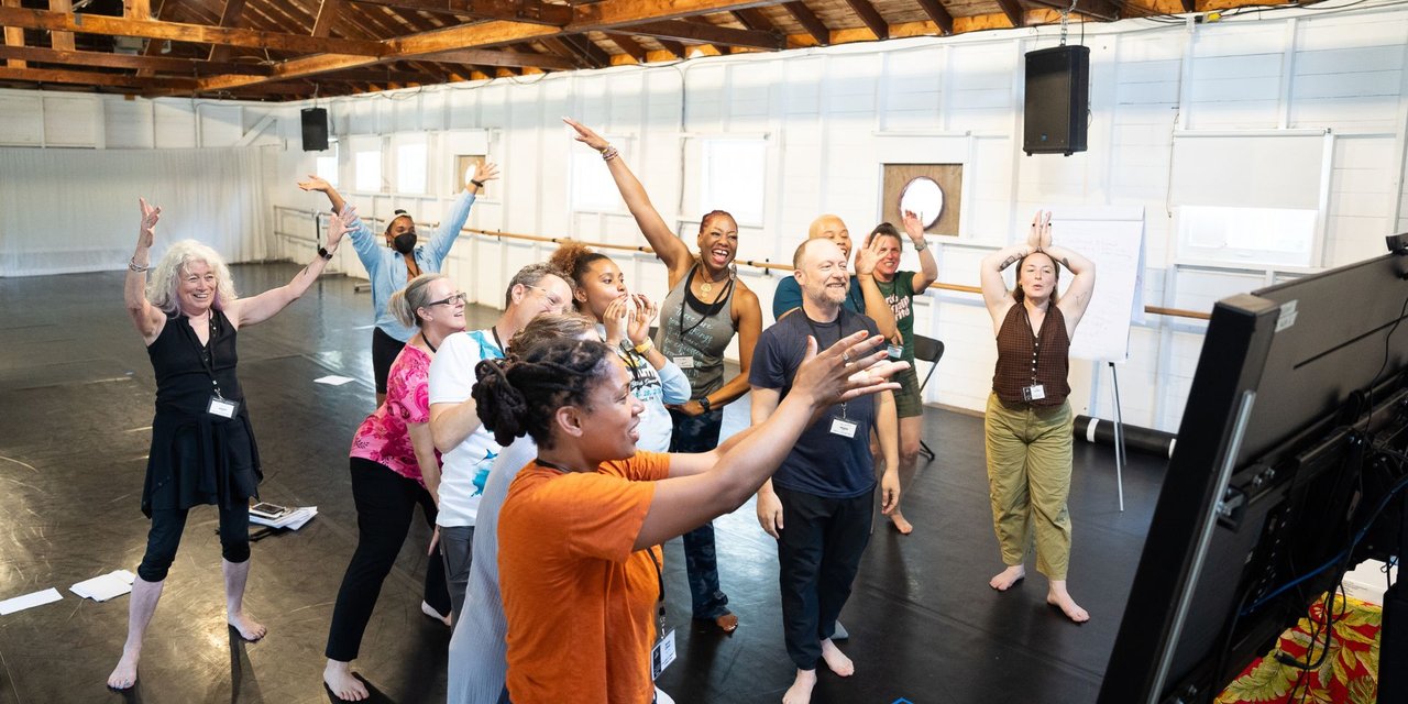 Eleven people of various ages, genders, and races stand in a studio facing computer monitor in the foreground. The group is expressing excitement by raising their arms at different levels.