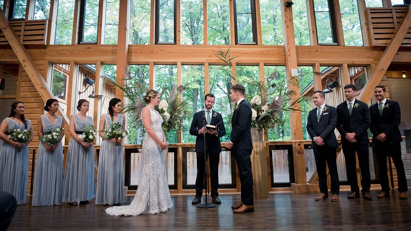 A bride and groom stand in front of an officiant with their wedding party behind them. They are in a dance studio with tall, bright windows showing lush trees behind them.