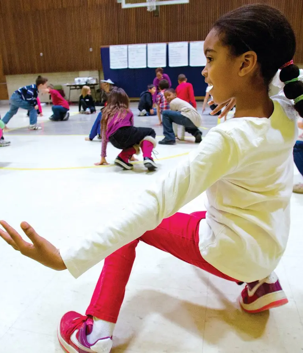 In a room full of children, one girl crouches close to the floor. Her left arm is stretched out to her side. She wears pink leggings and a white shirt.