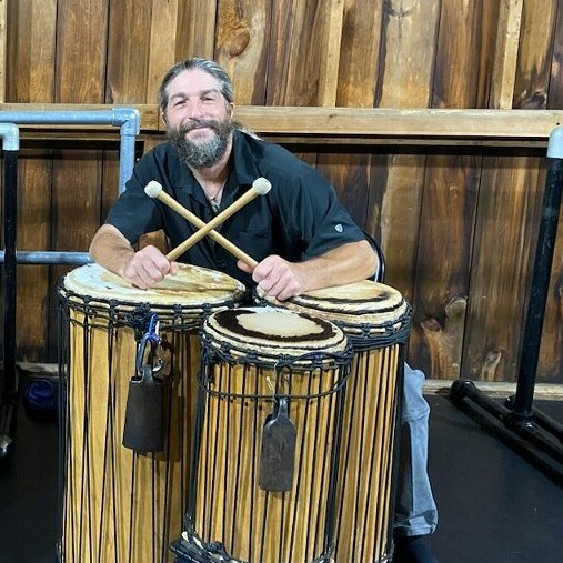 A man leans over three African drums, holding drumsticks crossed in front of him. He smiles for the camera.