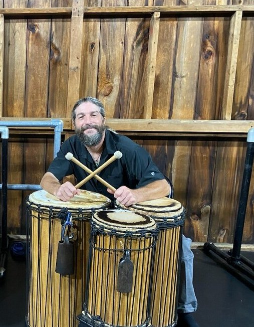 A man leans over three African drums, holding drumsticks crossed in front of him. He smiles for the camera.