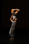 A male dancer lifts a female dancer above his head with her legs bent behind her.