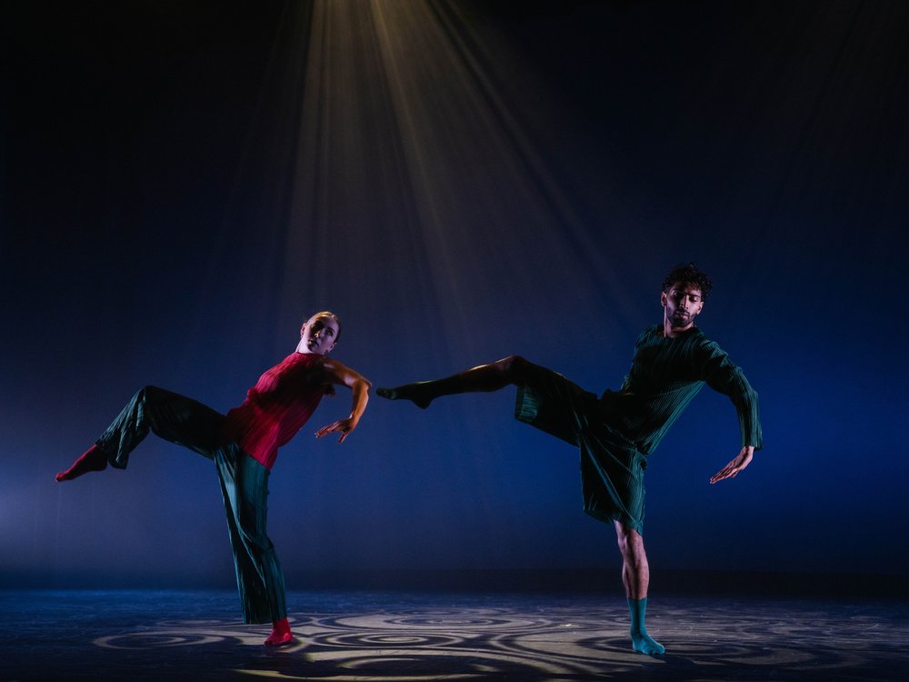 A female and male dance closely together, raising their right legs under stage lighting.