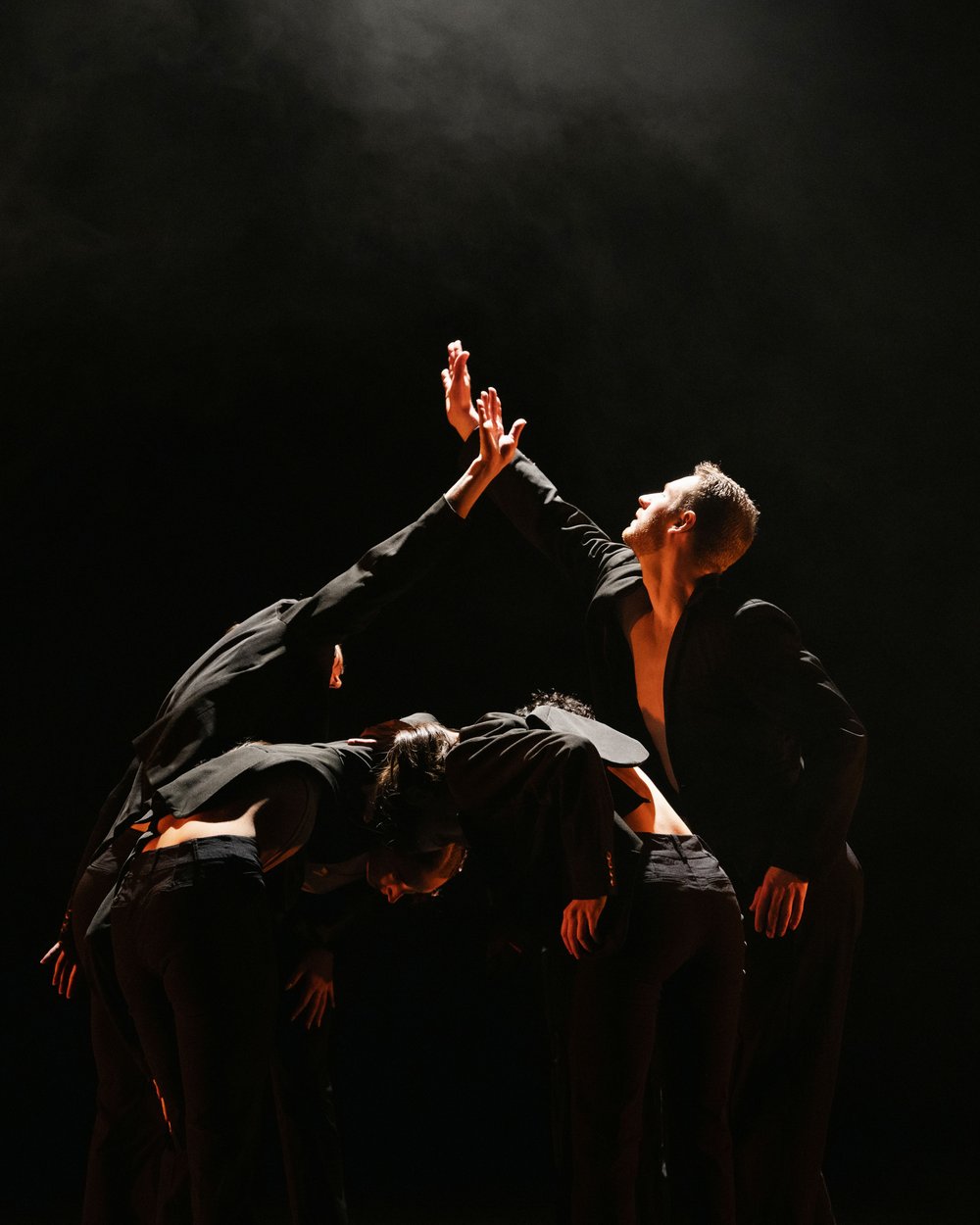 Five dancers wearing suits stand in a clump. Three dancers are bent forward in front of the clump with their heads together. The two dancers in the back stand facing each other with one arm raised, reaching for each other. They are illuminated by a single