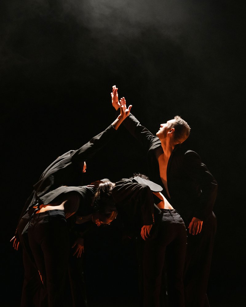 Five dancers wearing suits stand in a clump. Three dancers are bent forward in front of the clump with their heads together. The two dancers in the back stand facing each other with one arm raised, reaching for each other. They are illuminated by a single