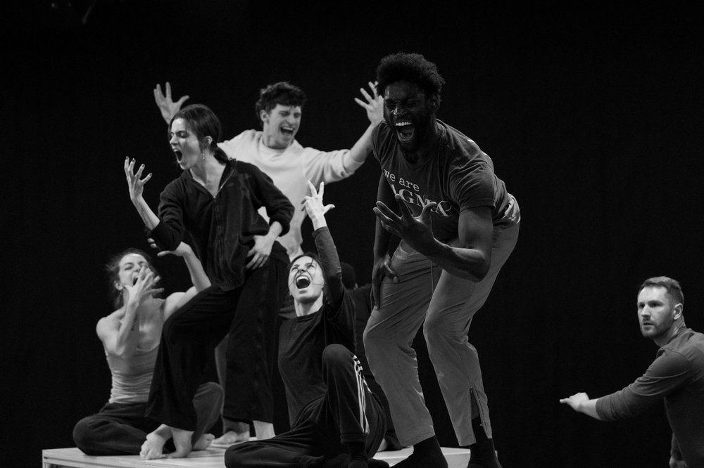 Six dancers sit or stand in formation on a long box-shaped prop in a dance studio. They all have their hands lifted in a claw shape in front of their faces, and have their mouths open and eyes closed, as if they are screaming.