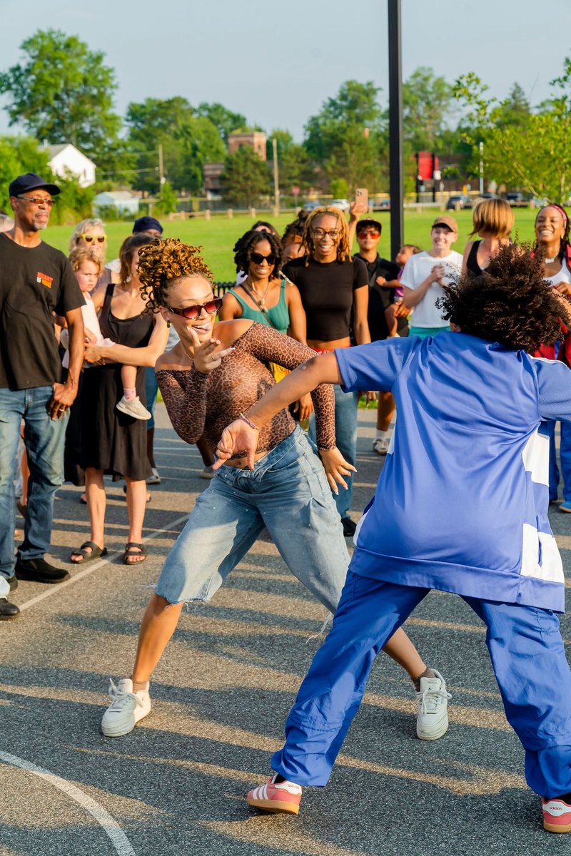 Two dancers face each other closely on an outdoor court while audience members crowd around them.
