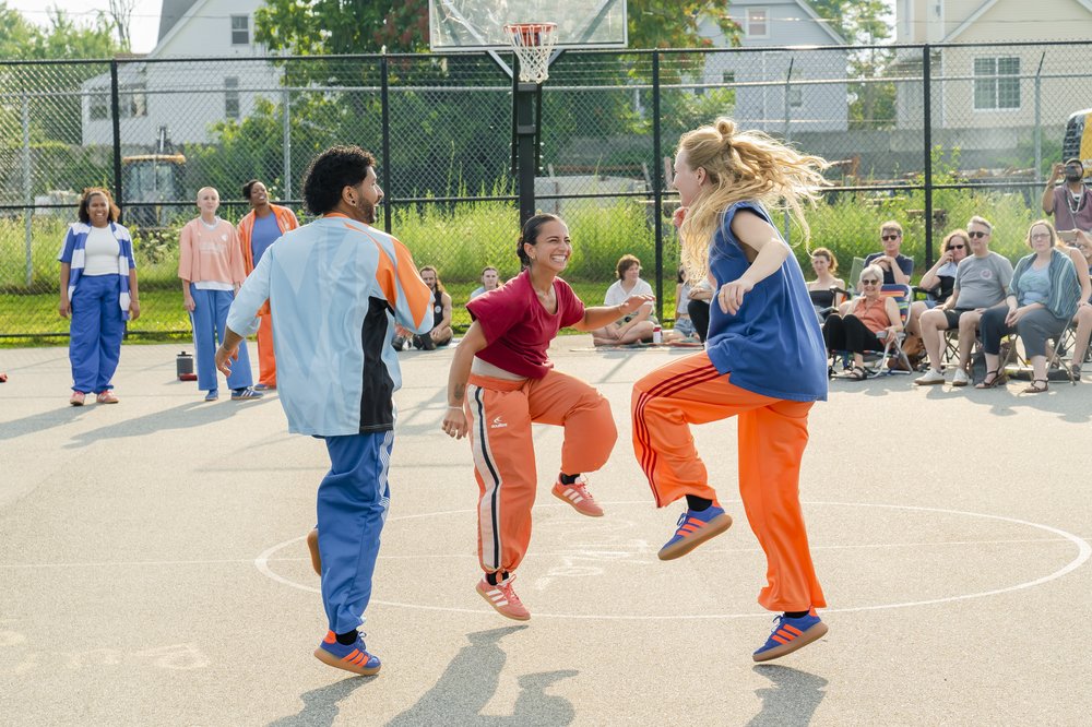 Three dancers hop simultaneously on an outdoor basketball court as audience members watch from behind.