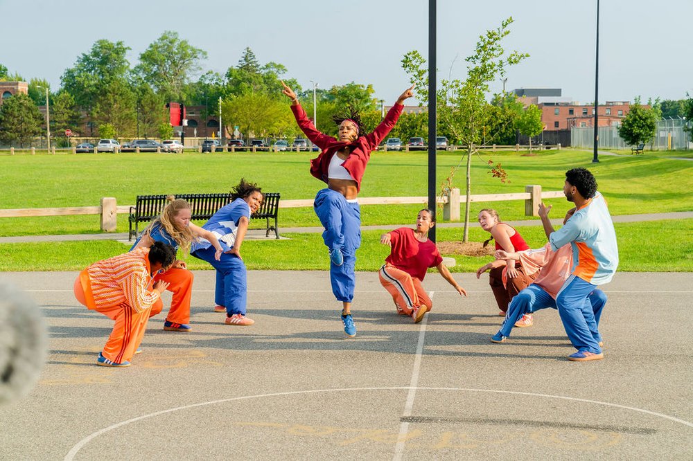 A group of dancers performs in formation on an outdoor basketball court with one dancer jumping in the center.
