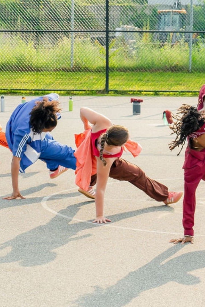 Dancers perform floor movement on an outdoor basketball court with seated audience members nearby.