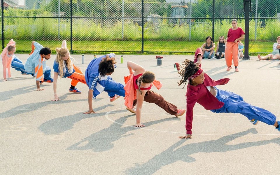 Dancers perform floor movement on an outdoor basketball court with seated audience members nearby.