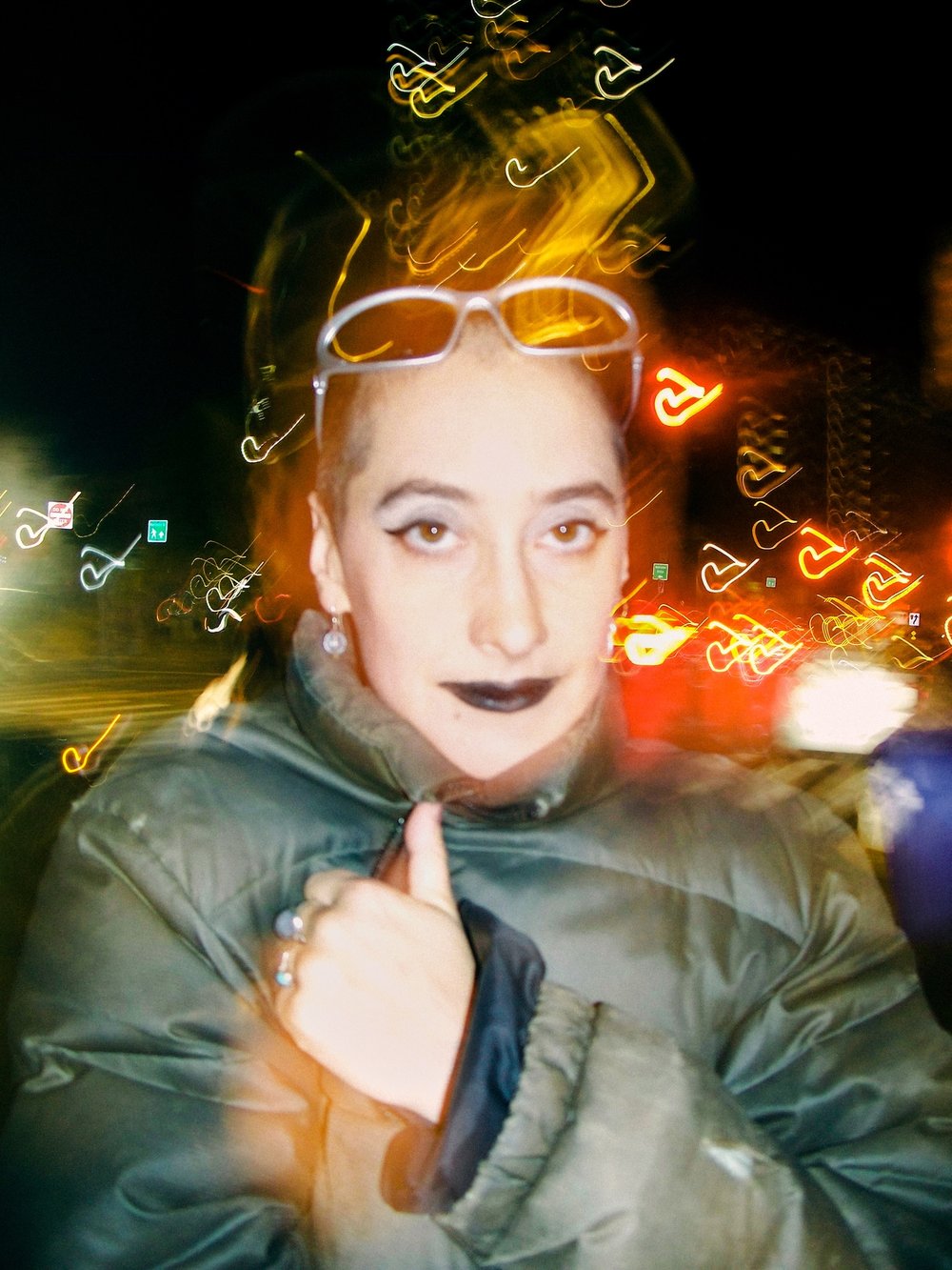 Liberty Styles is an androgynous person with a shaved head photographed at night on a city street. They are wearing black lipstick and hugging themselves in a big puffy coat.