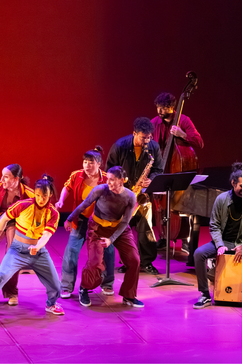 Seven dancers dressed in white, yellow, maroon, and gray dance on a stage with a purple lit floor and red lit background. The are in motion, taking a large step to their right all together. A small band plays next to them.