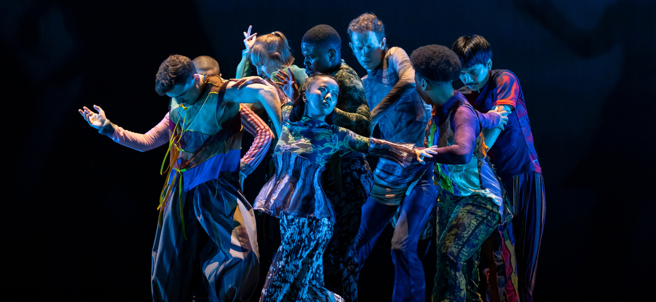 Eight dancers in colorful costumes stand closely together on a dark stage, extending their arms and leaning in different directions under blue stage lighting.