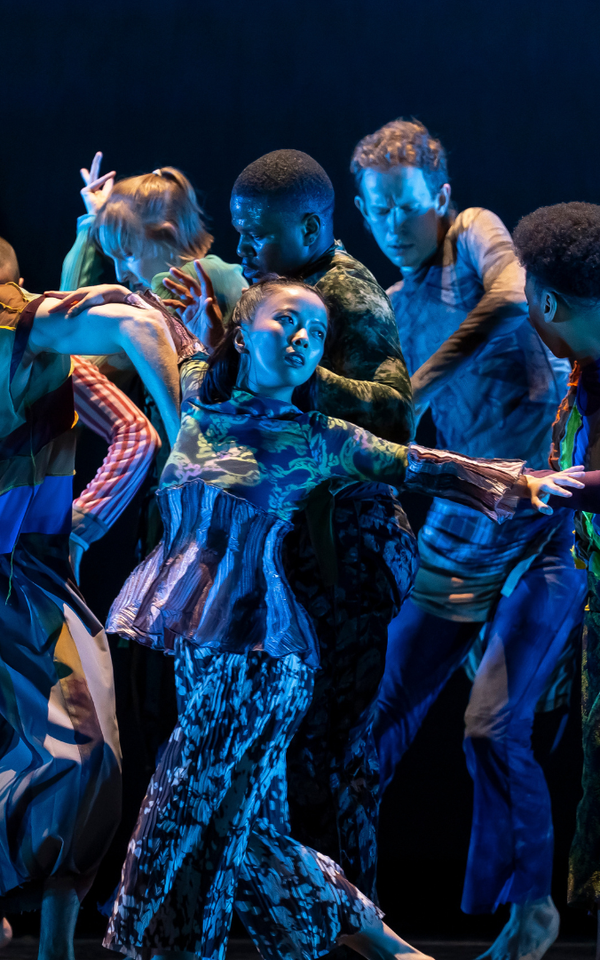 Eight dancers in colorful costumes stand closely together on a dark stage, extending their arms and leaning in different directions under blue stage lighting.
