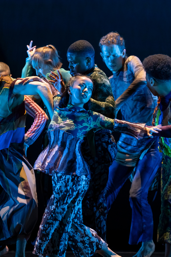 Eight dancers in colorful costumes stand closely together on a dark stage, extending their arms and leaning in different directions under blue stage lighting.